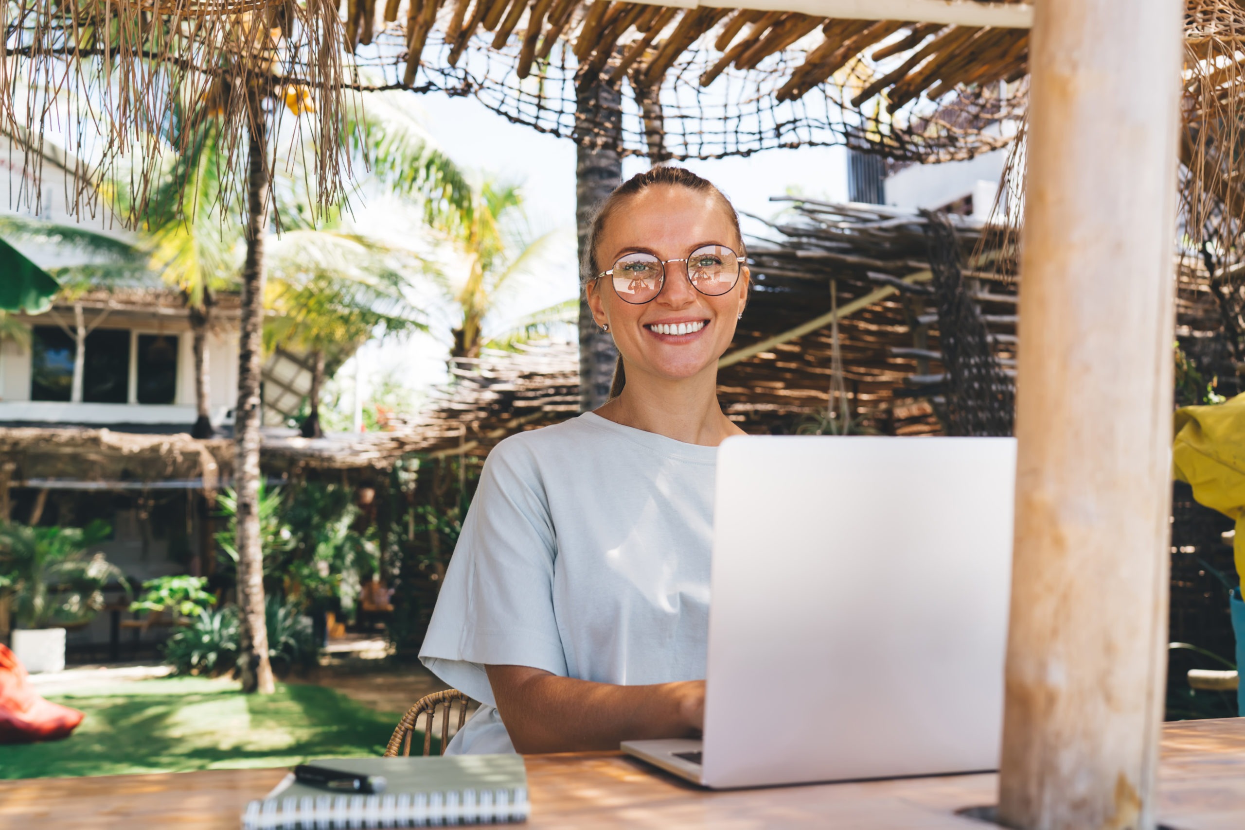 Positive young female freelancer in glasses and casual outfit working online on project using laptop sitting in gazebo near villa and palms