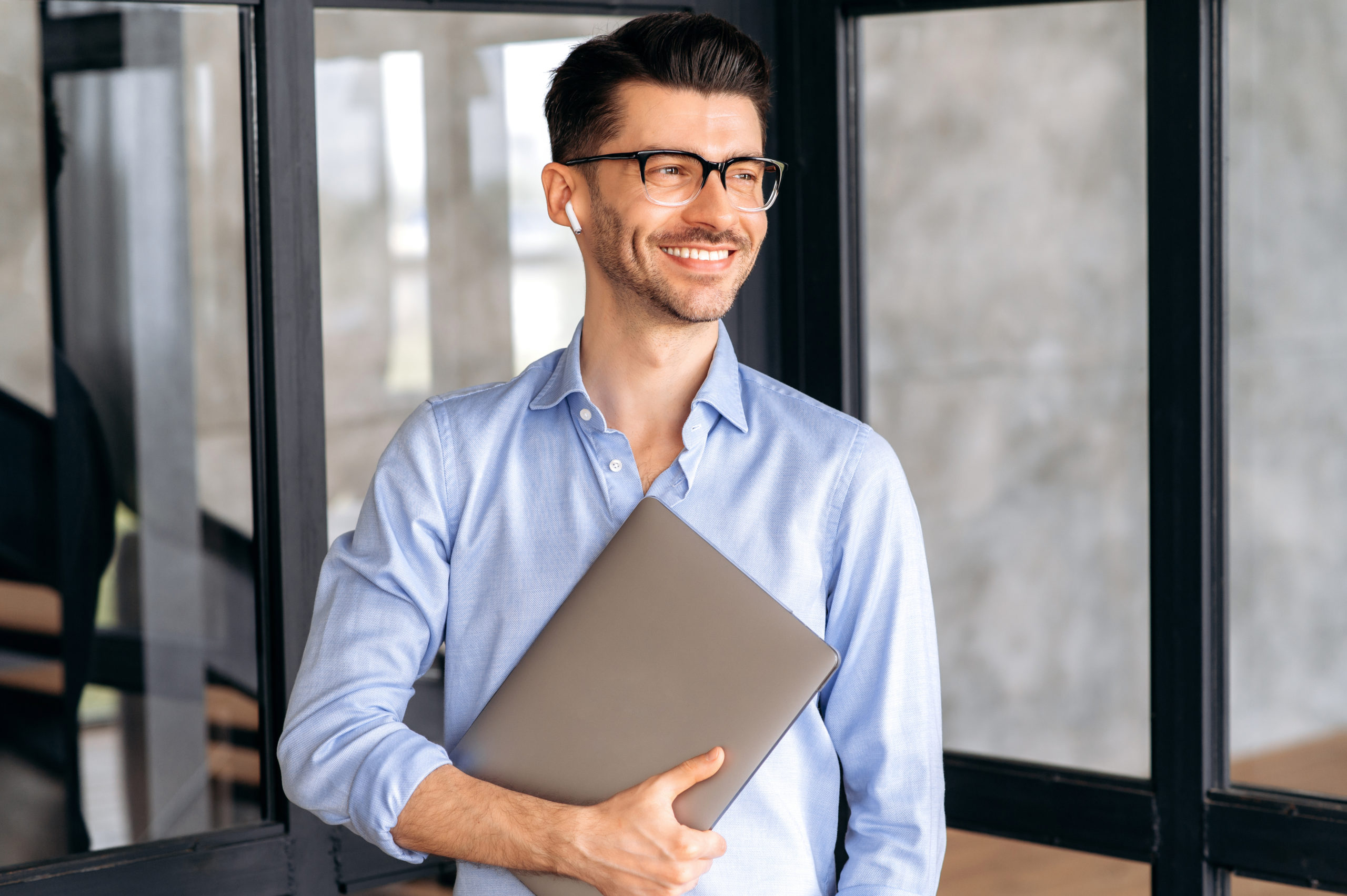 Portrait of a handsome confident successful caucasian businessman or office employee, wearing shirt and glasses, standing in office with laptop, looking to the side and smiling friendly