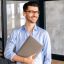 Portrait of a handsome confident successful caucasian businessman or office employee, wearing shirt and glasses, standing in office with laptop, looking to the side and smiling friendly